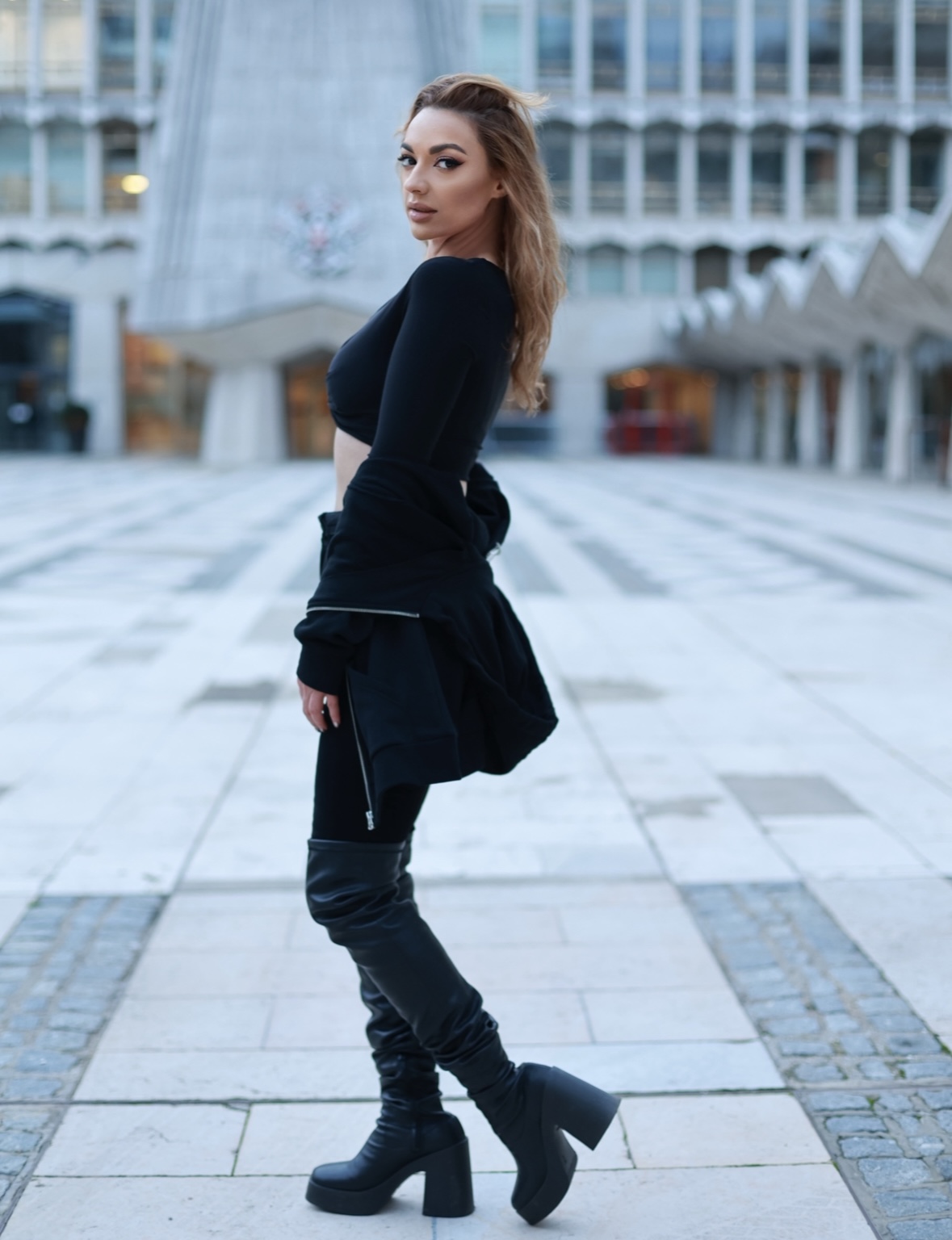 Chantelle is looking directly at camera with body facing to the side. stood in a paved courtyard wearing a black crop top, black jeans and boots